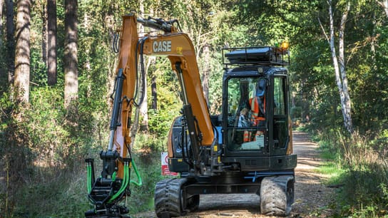 A compact excavator with a grading attachment works on a forest trail, surrounded by tall trees and dense greenery.