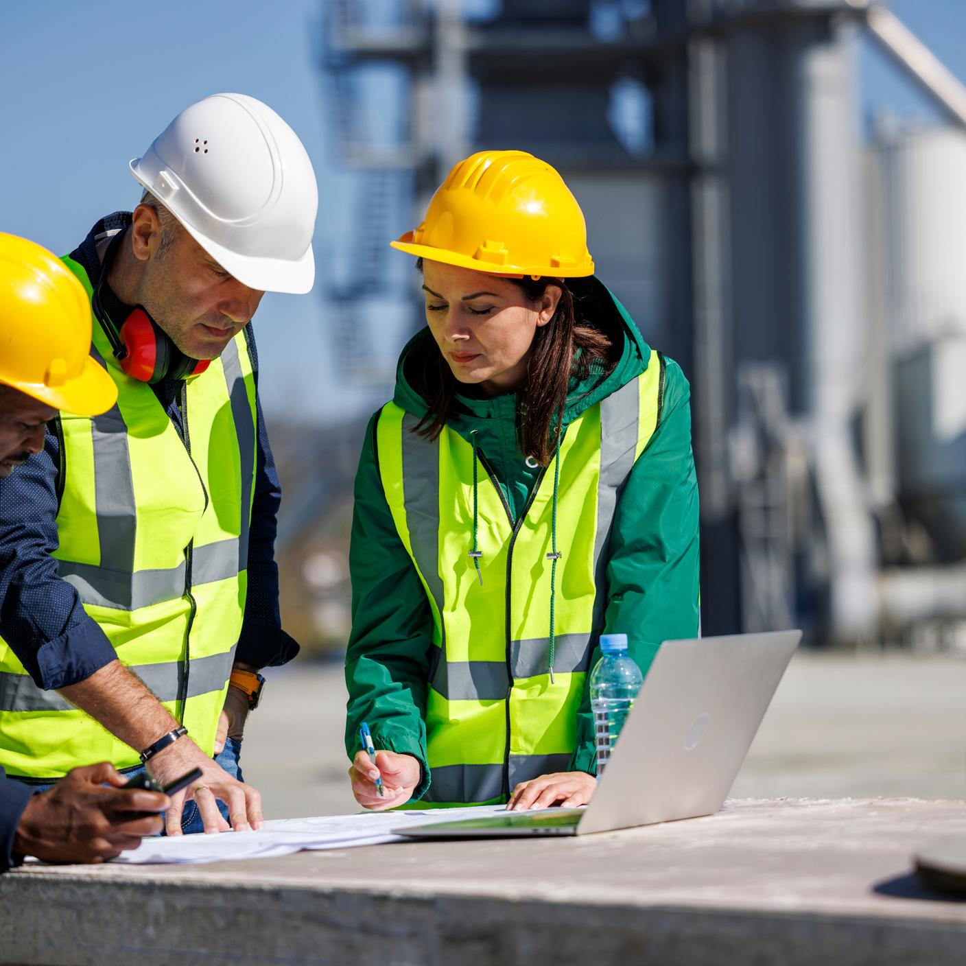 Three engineers in safety attire discussing at an industrial facility writing on paper next to a laptop.