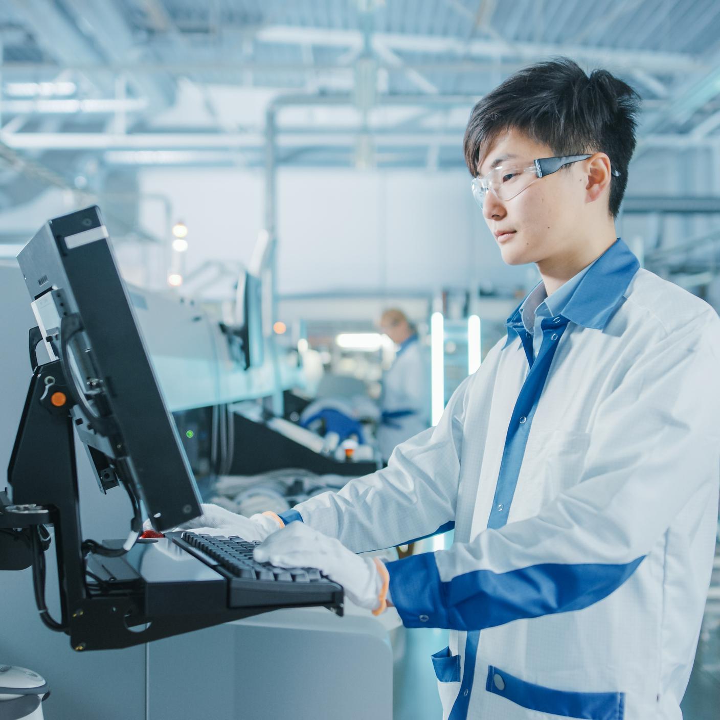 On High Tech Factory Asian Engineer Uses Computer for Programing Pick and Place Electronic Machinery for Printed Circuit Board Surface Mount Assembly Line.