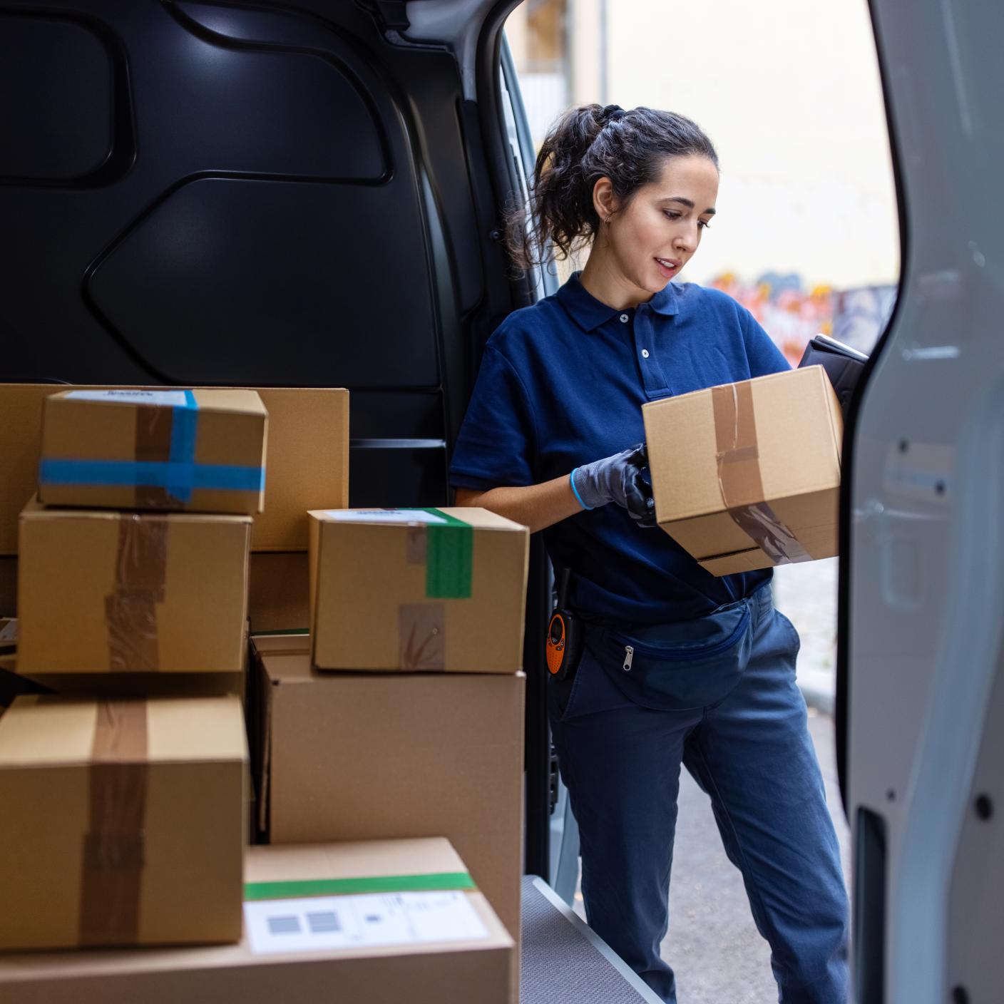 Female courier standing by shipping van checking the parcel for delivery. Woman looking at the delivery address of the parcel in van.