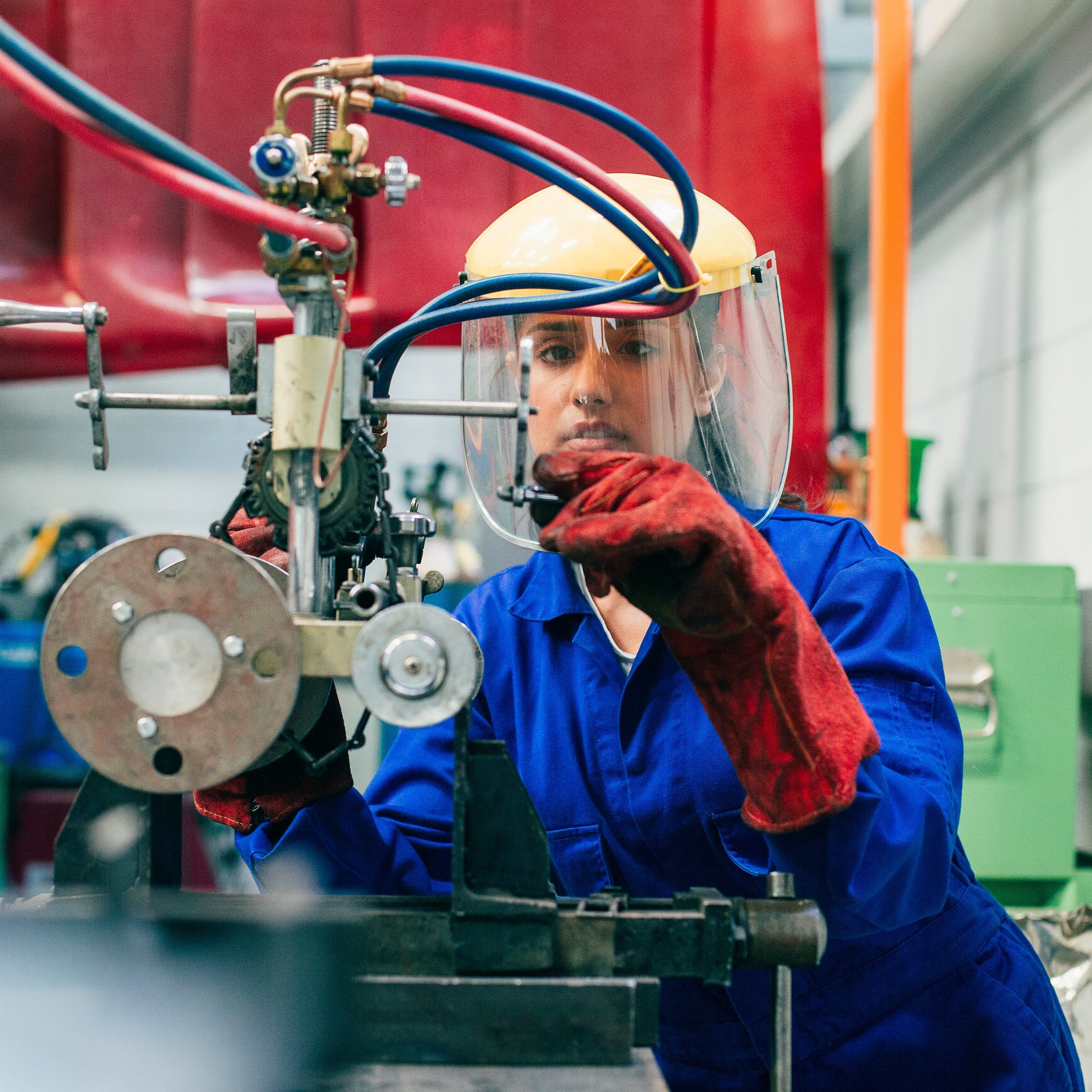 A young woman is using engineering equipment in a workshop. She is wearing a protective mask and gloves as well as blue coveralls.