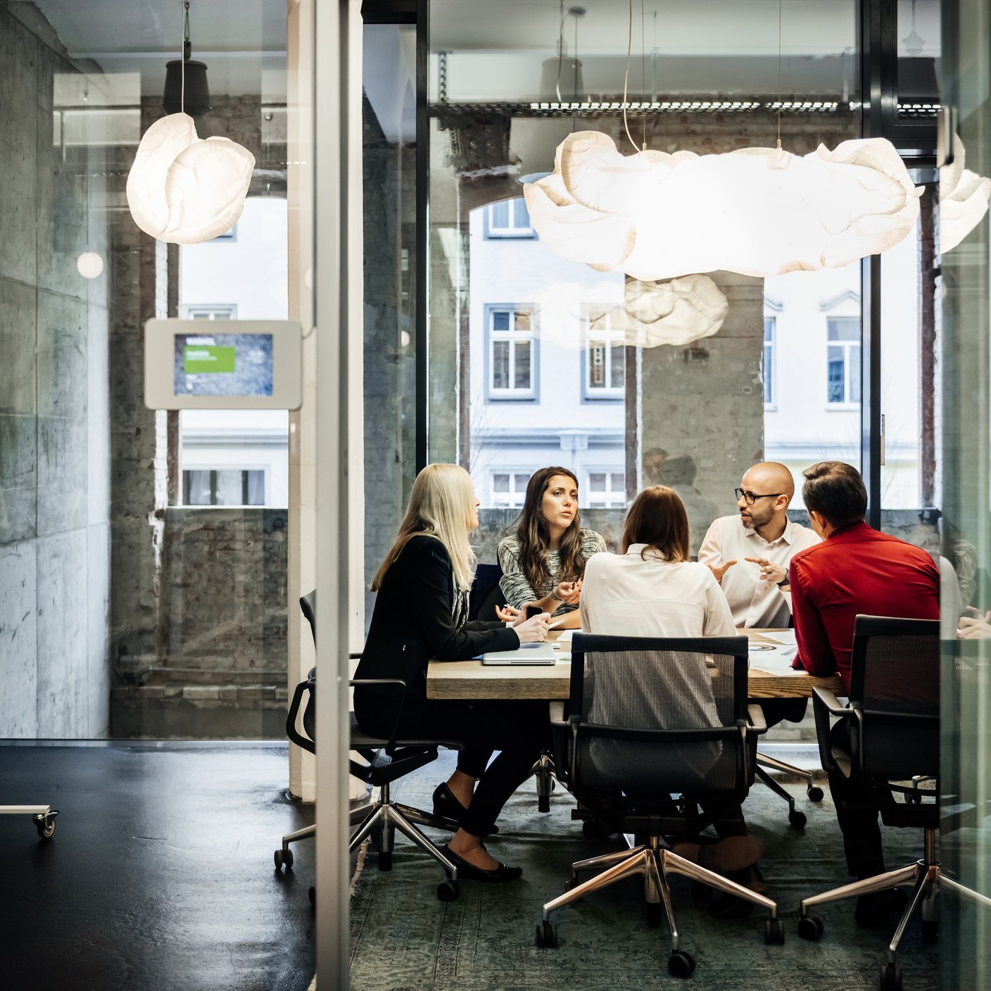 Business meeting in a modern office. A group of people is sitting at a table during a business meeting in a bright, modern office. The team is talking business while pie charts can be seen hanging on the wall. Big bright windows are seen in the background.