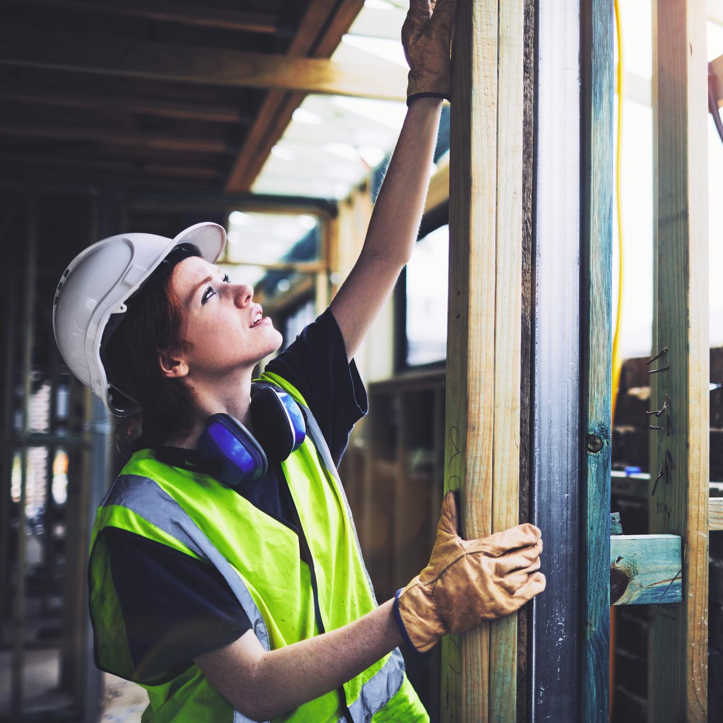 Construction worker wearing safety gear reaches up to adjust a wooden frame inside a partially built structure.