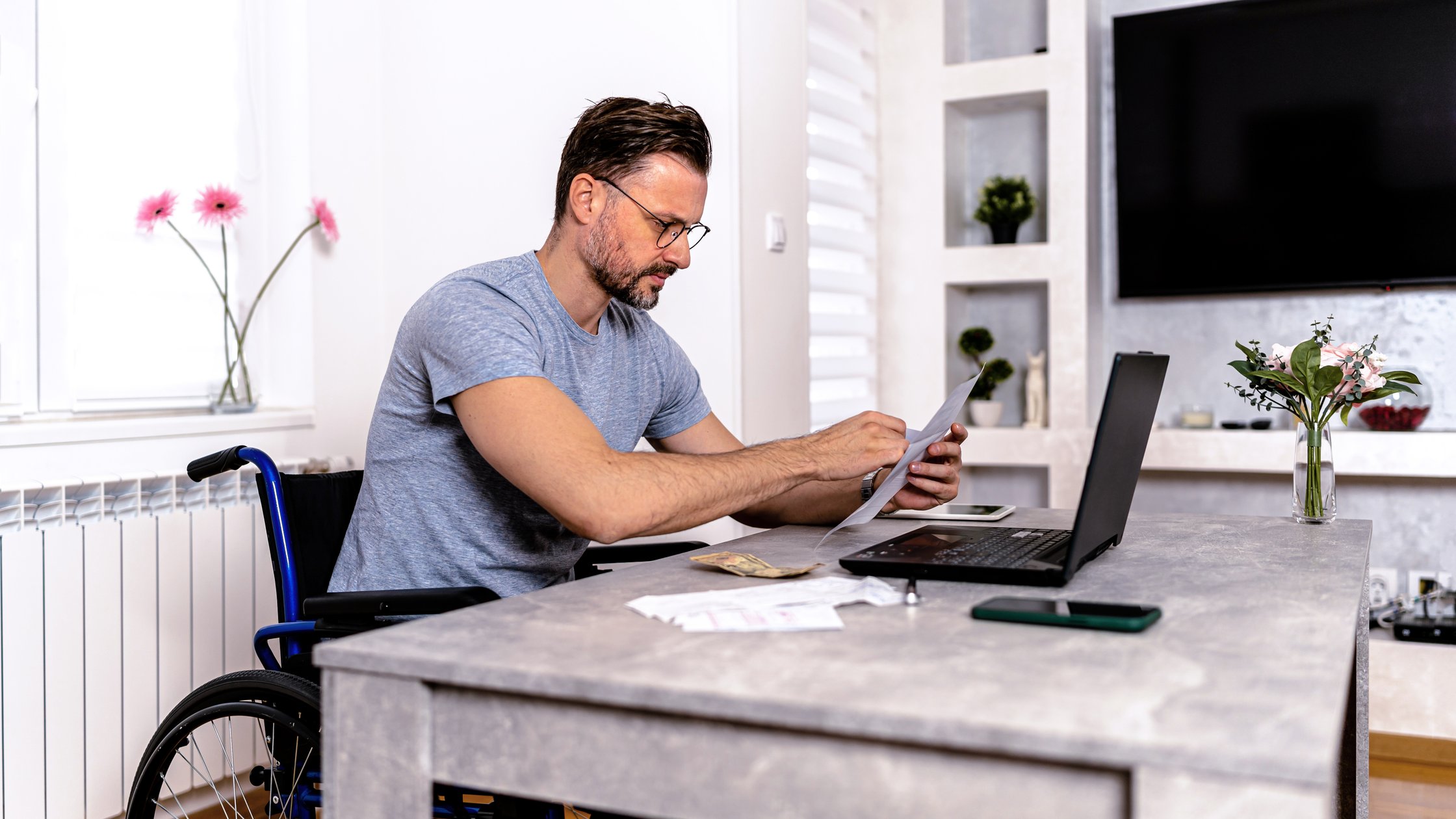 Using his wheelchair as a seat, a man is paying his bills online. He looks at his computer screen intently while his fingers click away on the keyboard