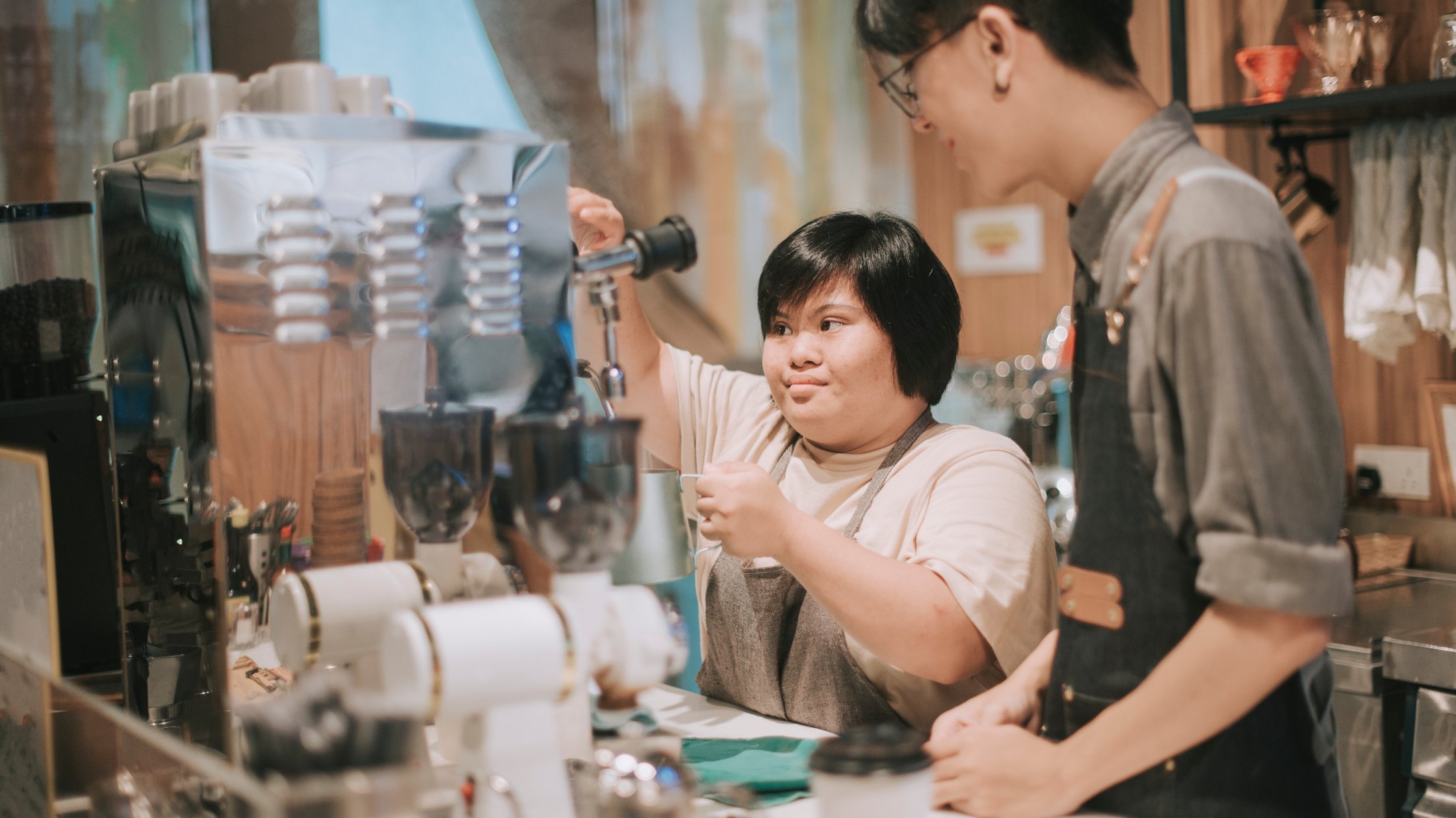 Asian Down Syndrome female learning from barista making coffee in her café place of work.